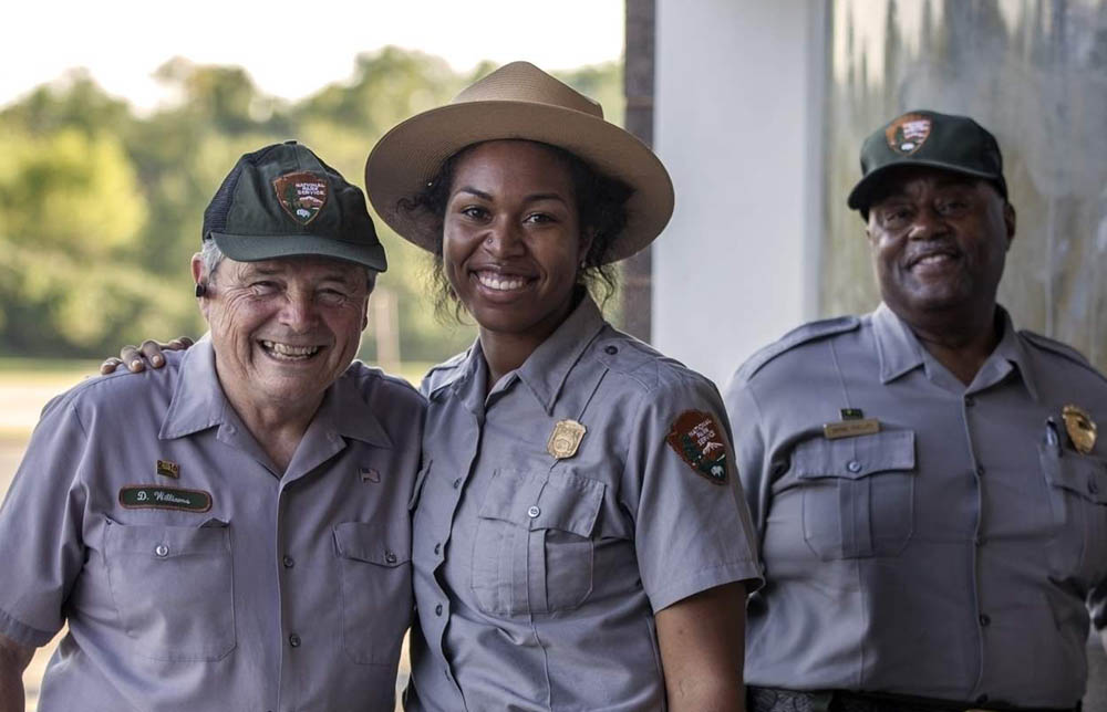 Three people stand in NPS uniforms. Only two wear badges. A woman wears a gold shield shaped badge and a man a large gold arrowhead shaped badge.